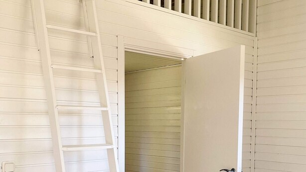 Un interior en tonos blancos con puerta de madera, una escalera blanca y un techo inclinado.