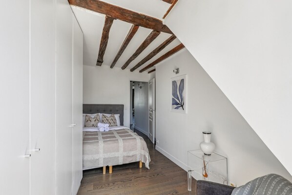 A bedroom with a wooden ceiling, a bed with a patterned bedspread, a bedside table with a lamp, and a door leading to another room.