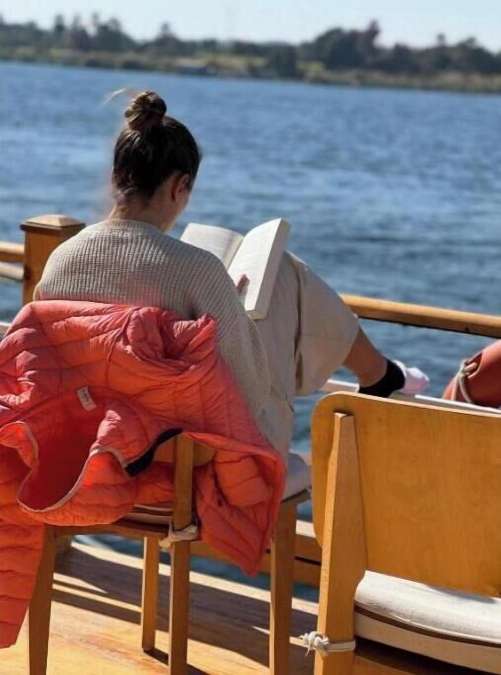 A person sitting on a wooden bench by a body of water, reading a book.