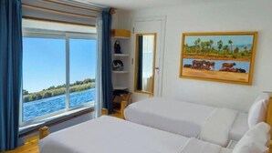 Una habitación con dos camas, un ventanal con vista al mar, un cuadro de vacas en una playa y suelo de madera.