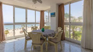 A room with a dining table and chairs, large windows with curtains, a ceiling fan, and a view of the beach. - Apartamento Joya 3 by Costa CarpeDiem (Calpe)