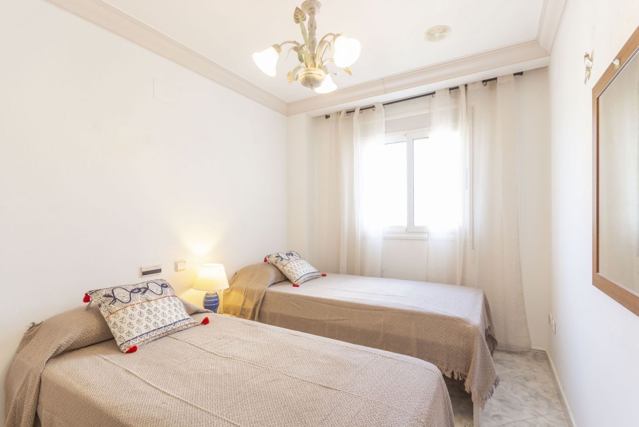 A hotel room with two beds, a chandelier, a window with curtains, and a nightstand with a lamp.