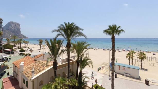 A beach view with palm trees, a building with a red roof, and a clear blue sky. - Apartamento Joya 3 by Costa CarpeDiem (Calpe)