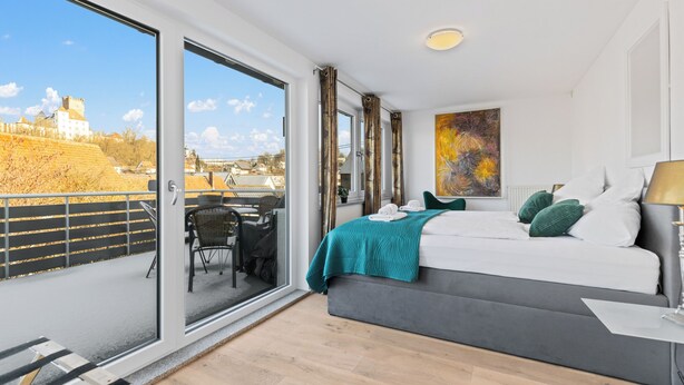 Una camera d'albergo moderna con un grande letto, vista su un castello dal balcone, e un'area pranzo con tavolo e sedie.