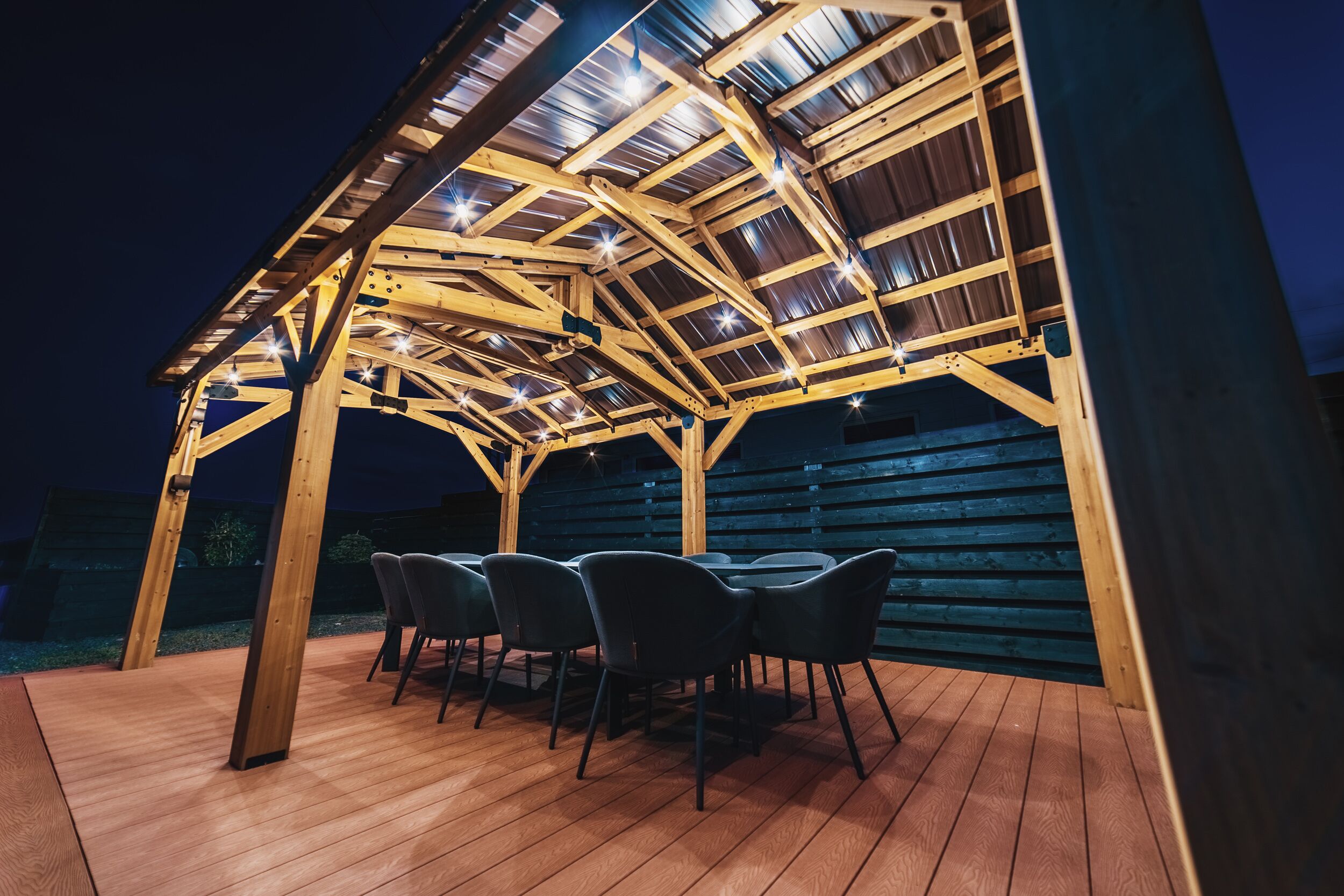 A wooden pergola structure with a roof supported by beams and string lights, surrounded by a wooden deck and four chairs.