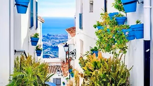 A narrow street with white buildings, blue hanging planters, and a view of the sea.