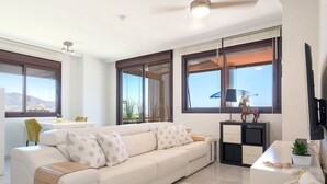 A modern living room with a sofa, a coffee table, and a TV stand. There are large windows with a view of the outdoors.