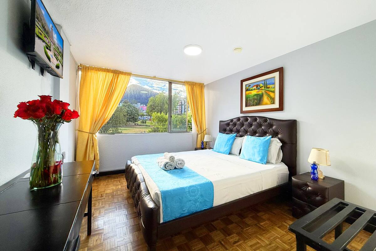 Habitación de hotel con cama, escritorio, silla, jarrón de flores, ventana con cortinas y un cuadro en la pared.