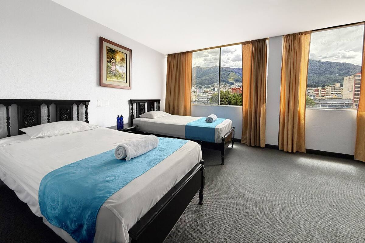 Habitación de hotel con dos camas, un ventanal con cortinas, un cuadro en la pared y vistas a la ciudad.