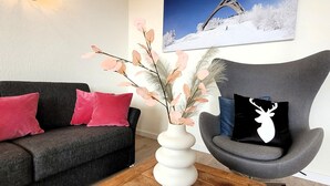 A living room with a dark grey sofa, red cushions, a white vase with branches, a wooden coffee table, and a grey armchair with a black cushion featuring a deer silhouette. - Apartment in the Centre of Winterberg (Winterberg)