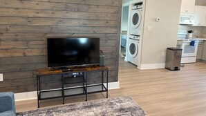Ein modernes Wohnzimmer mit einem Flachbildfernseher auf einem Holzständer, eine Küche mit weißen Schränken, einer Waschmaschine und einem Edelstahl-Mülleimer.
