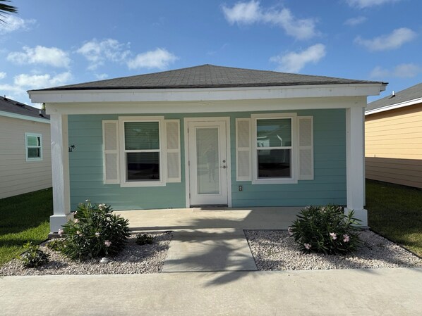 A single-story house with a front door, two windows with shutters, and a small porch area with plants.