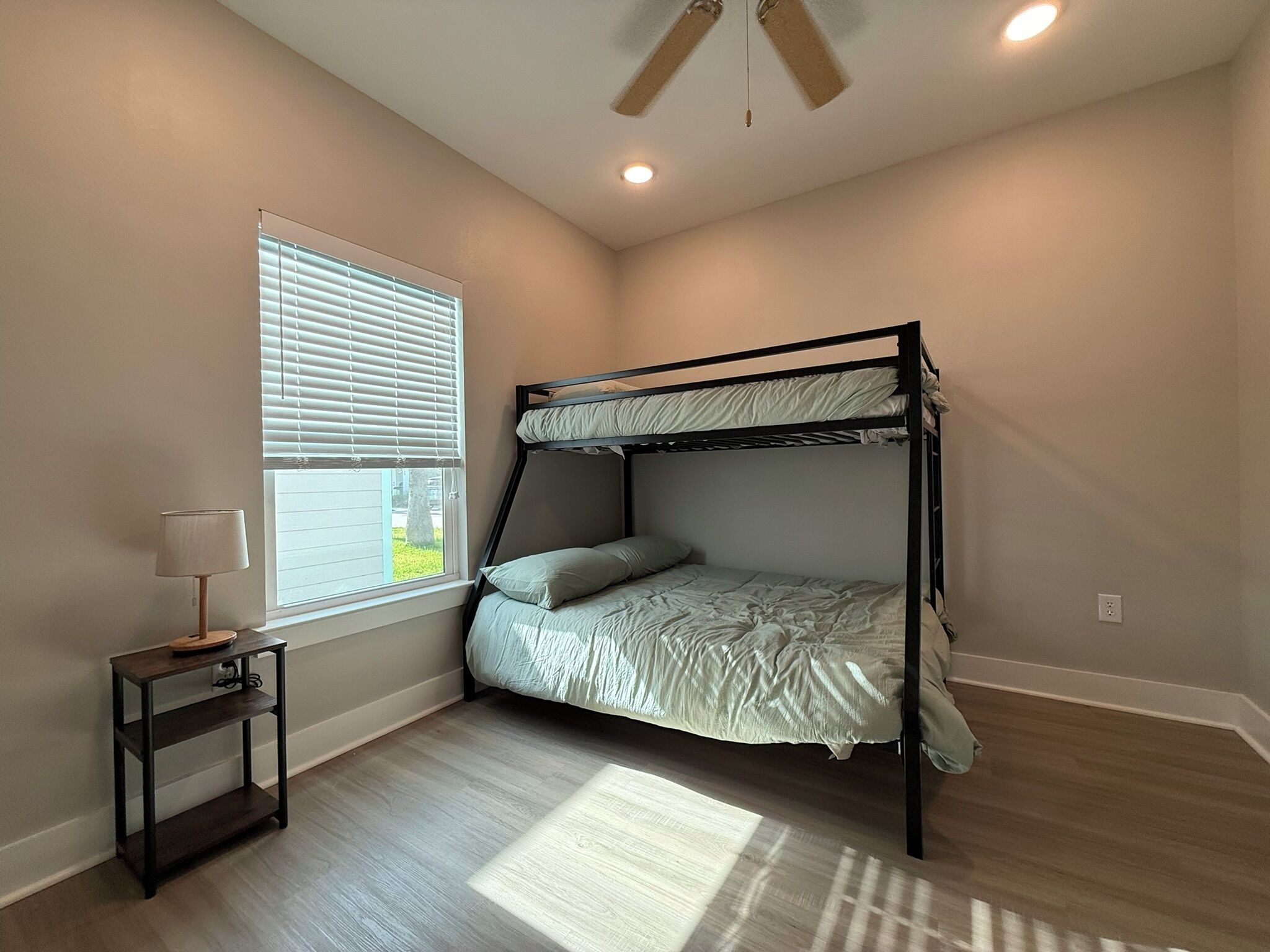 A bedroom with a bunk bed, a small table with a lamp, a window with blinds, and a ceiling fan.