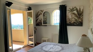 A bedroom with a bed, a nightstand, a lamp, a window with curtains, a mirror, and a view of the outside. - MAGNIFICENT VILLA SEA VIEW 4 BEDROOMS SWIMMING POOL QUIET ON THE HEIGHTS OF BORMES (Bormes-les-Mimosas)