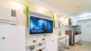 A modern kitchen with a built-in TV displaying 'BLACK SUMMER', white cabinetry, a stainless steel refrigerator, and a small bar area with stools. - Infini by J&J ( City Center |Near Malls ) (Cebu City)