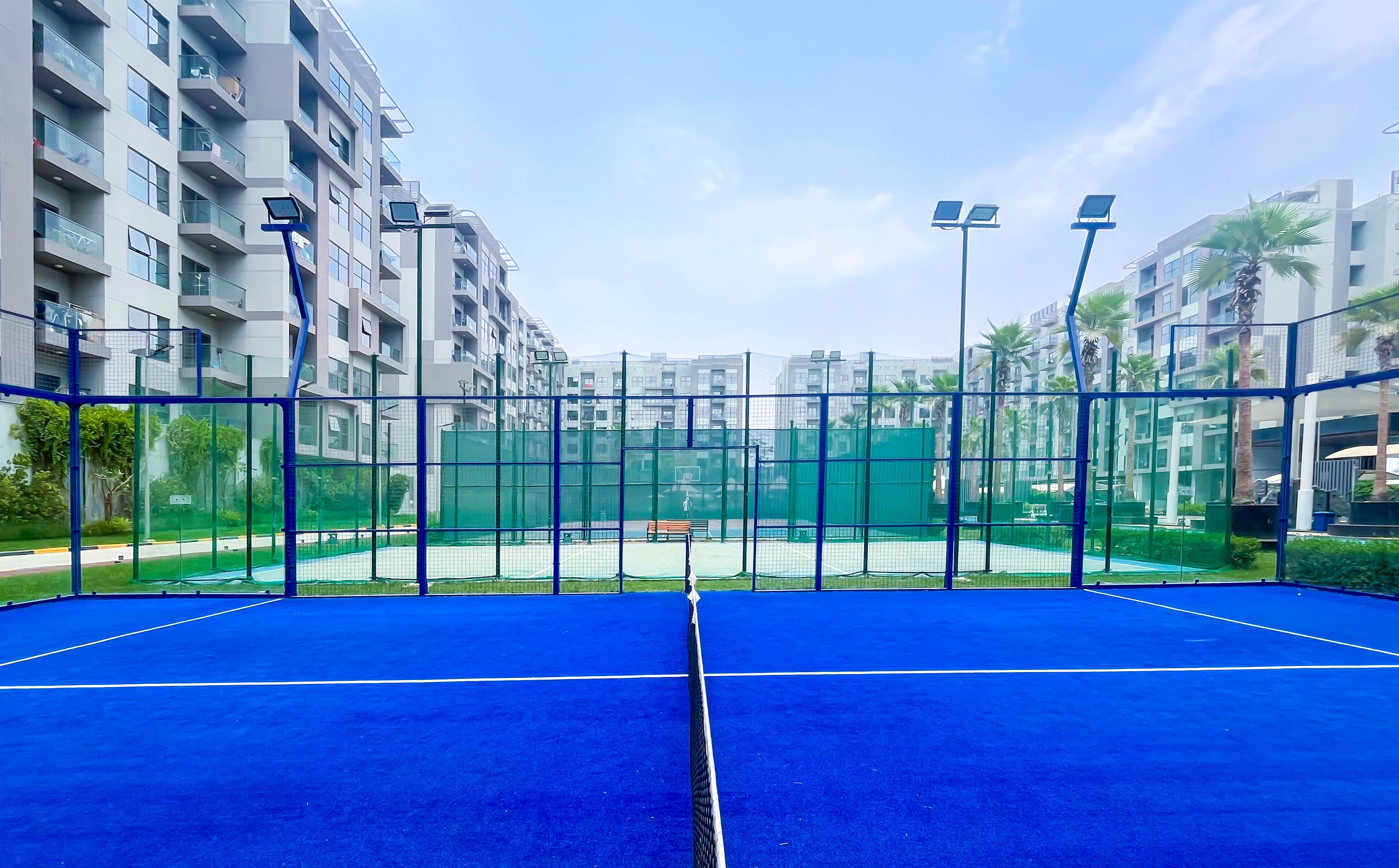 An indoor tennis court with a blue surface, surrounded by a green fence, and adjacent to residential buildings.