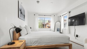 A modern bedroom with a large bed, a bedside table with a lamp, a TV mounted on the wall, and a window with a view of a building. - Pool & Hot Tubs | Nextdoor to Pickleball | Air Conditioning (Steamboat Springs)