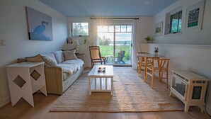 A cozy living room with a sofa, coffee table, and chairs, a fireplace, and a large window with a view of greenery. - Coastal Blue Heron Apartment at Tapley Farm, Blue Hill Peninsula, Acadia (Brooksville)