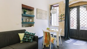 A modern living room with a sofa, a dining table with chairs, a window with curtains, and a wall-mounted shelf with decorative items. - Studio _ large openspace with kitchen and courtyard (Siracusa)