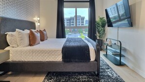 Un dormitorio moderno con cama, ventilador de techo, televisor y vistas a la ciudad a través de amplias ventanas.
