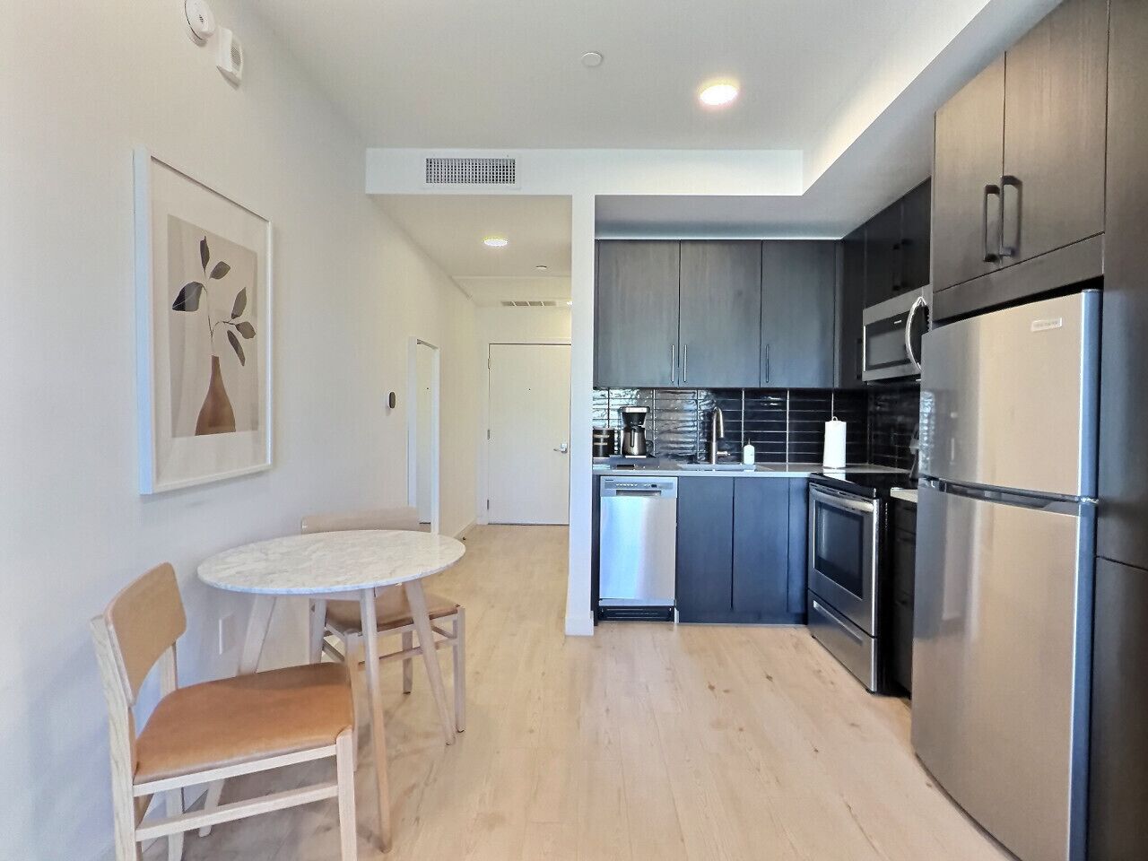 A modern kitchen with stainless steel appliances, a dining area with a round table and chairs, and a hallway leading to a door.