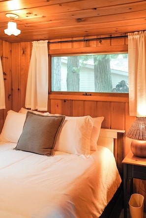 Une pièce de cabane en bois comprenant un lit, des rideaux blancs, une fenêtre et une lampe.
