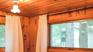 Ein Zimmer in einer Holzhütte mit einem Bett, weißen Vorhängen und einem Fenster mit Blick auf Bäume.