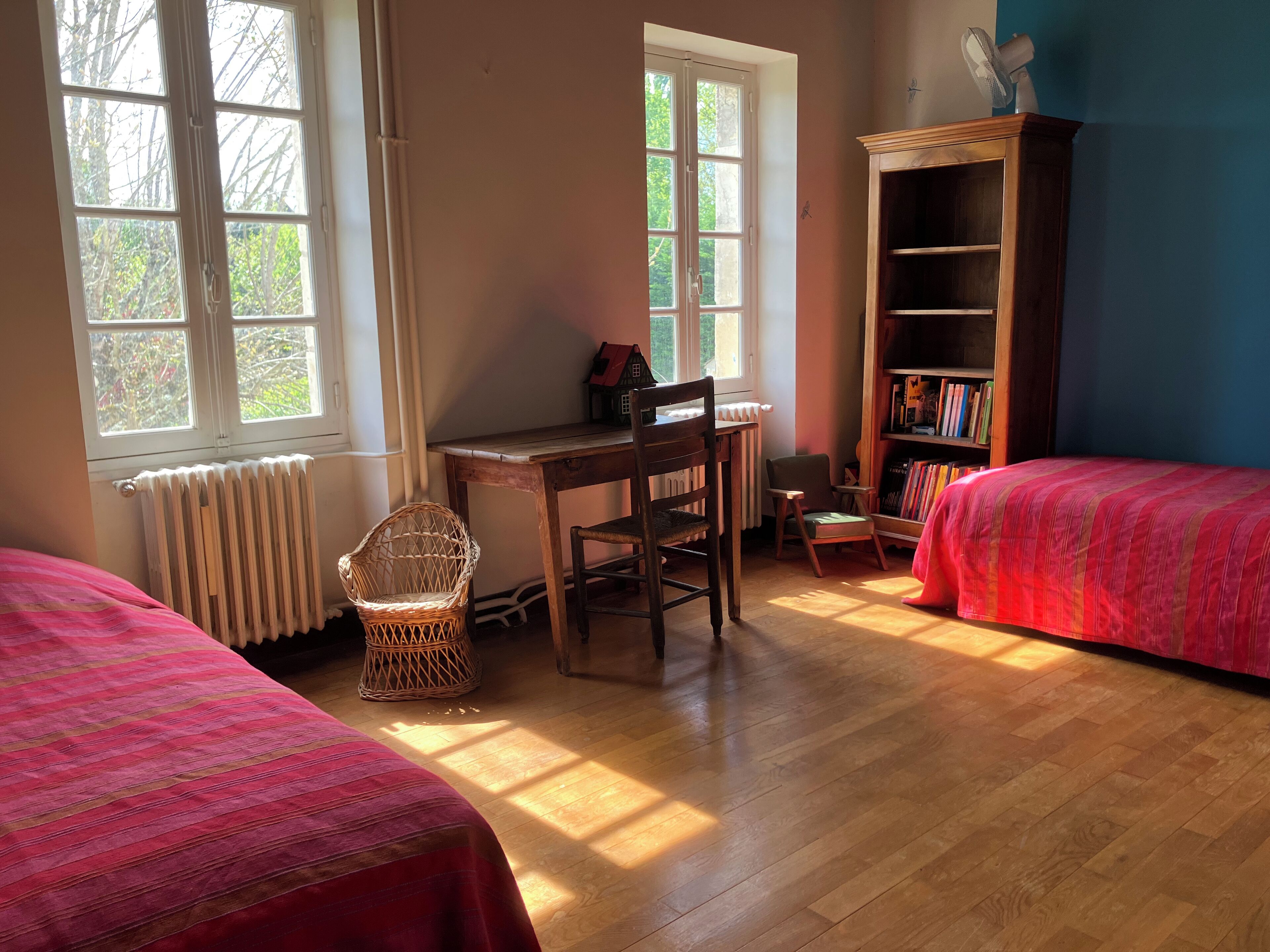 Une pièce comprenant un bureau en bois, deux chaises, un fauteuil en osier, une bibliothèque et deux lits avec une literie rouge.