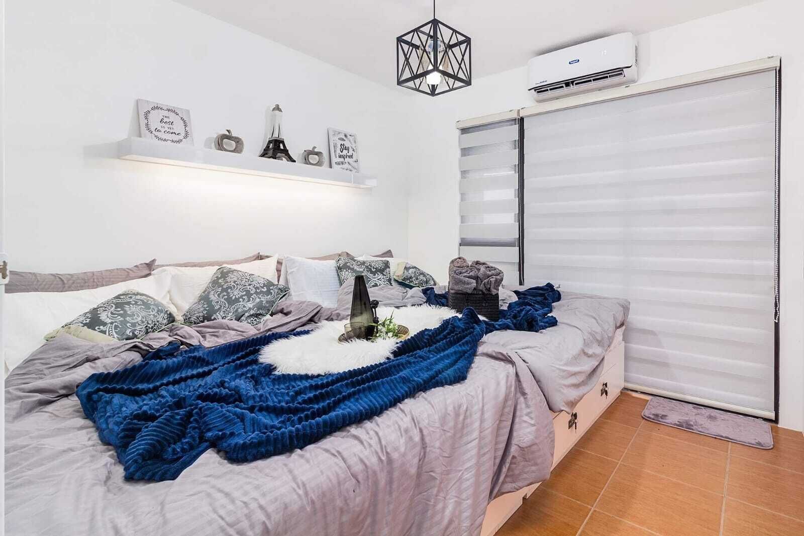 A bedroom with a bed, pillows, a blanket, a bedside table, a wall-mounted air conditioner, and a window with blinds.