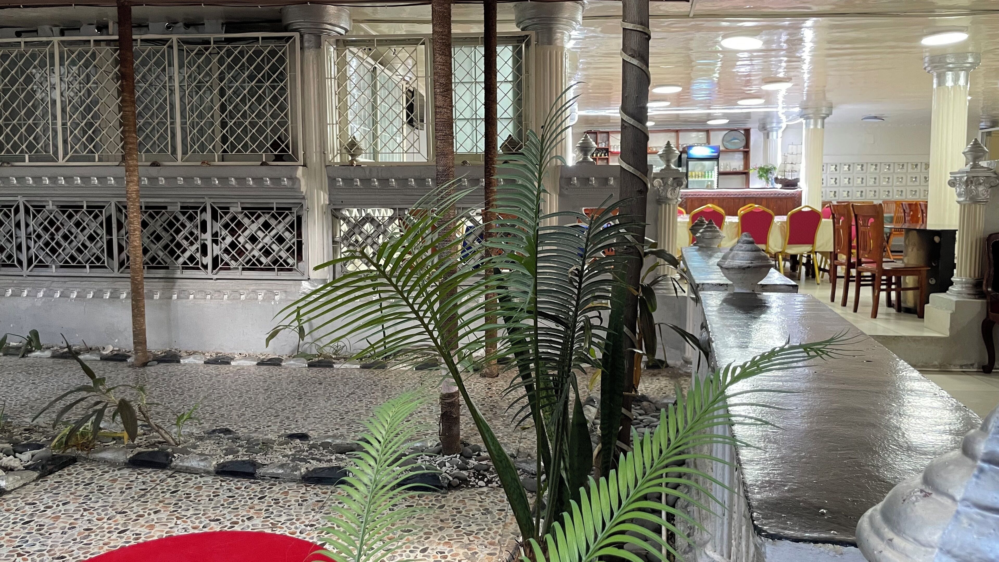 A hotel lobby with a tiled floor, palm trees, a red carpet, and a long table with chairs.