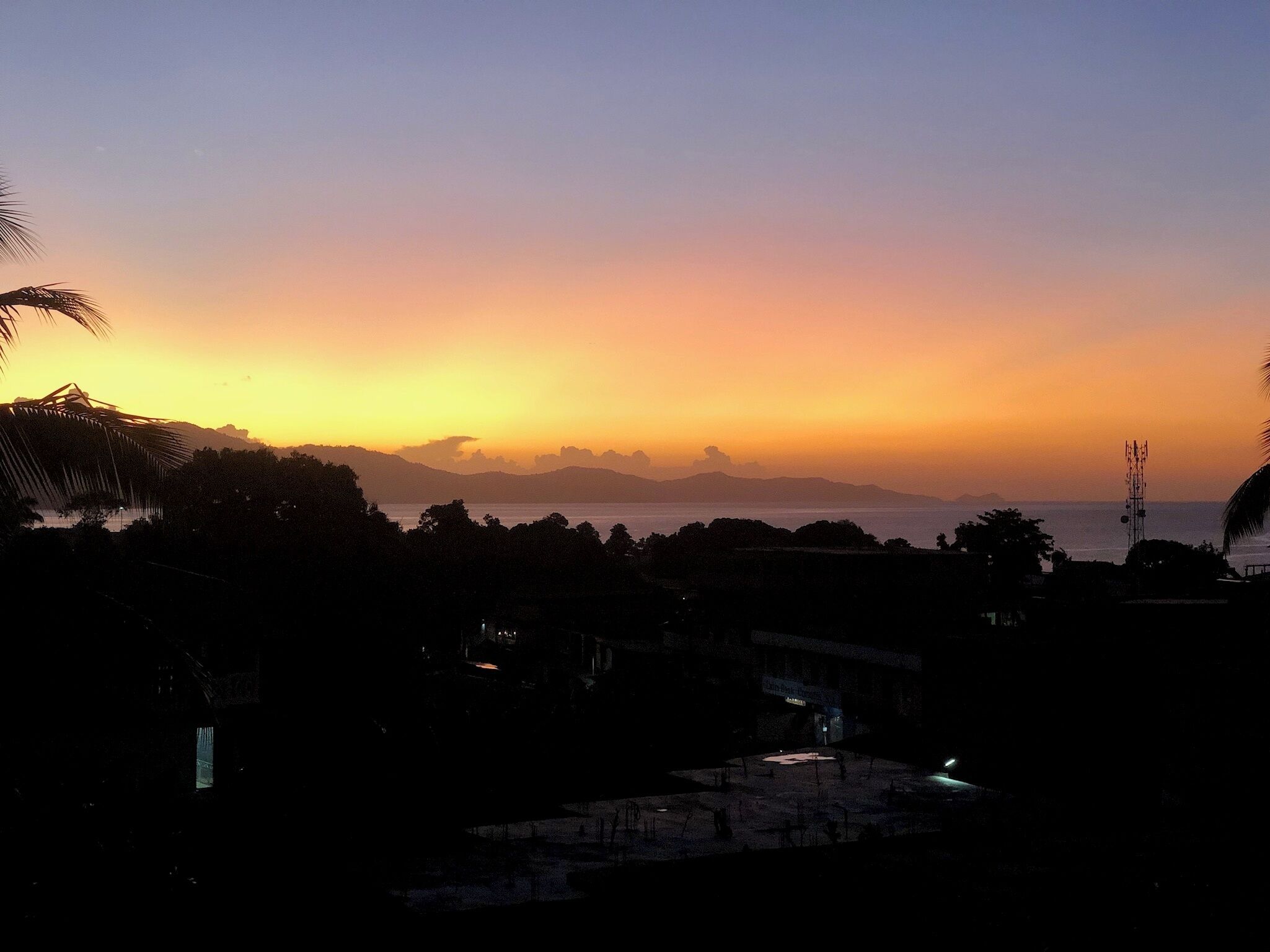 Una vista del atardecer costero con siluetas de árboles y una lejana cadena montañosa.