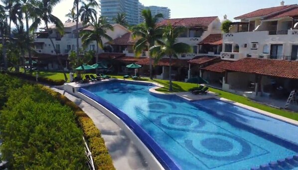 A swimming pool with a geometric pattern, surrounded by palm trees and a building with a red-tiled roof.