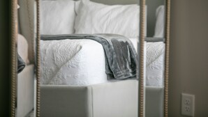 Una cama con colcha blanca y manta gris, un espejo con marco de madera, y una pared blanca con toma de corriente.