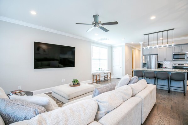 A modern living room with a large flat-screen TV, a ceiling fan, and a kitchen area with a stainless steel refrigerator and bar stools.