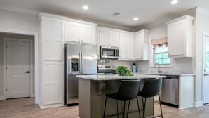Una cocina moderna con armarios blancos, un refrigerador de acero inoxidable, un microondas y un lavavajillas. Hay una pequeña isla con dos taburetes negros.