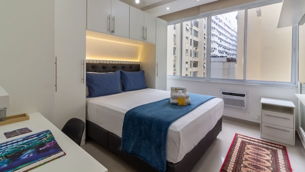 Una habitación de hotel moderna con una cama grande, un escritorio, una silla, un ventilador de techo, una ventana con vista a la ciudad y una mesita de noche pequeña.