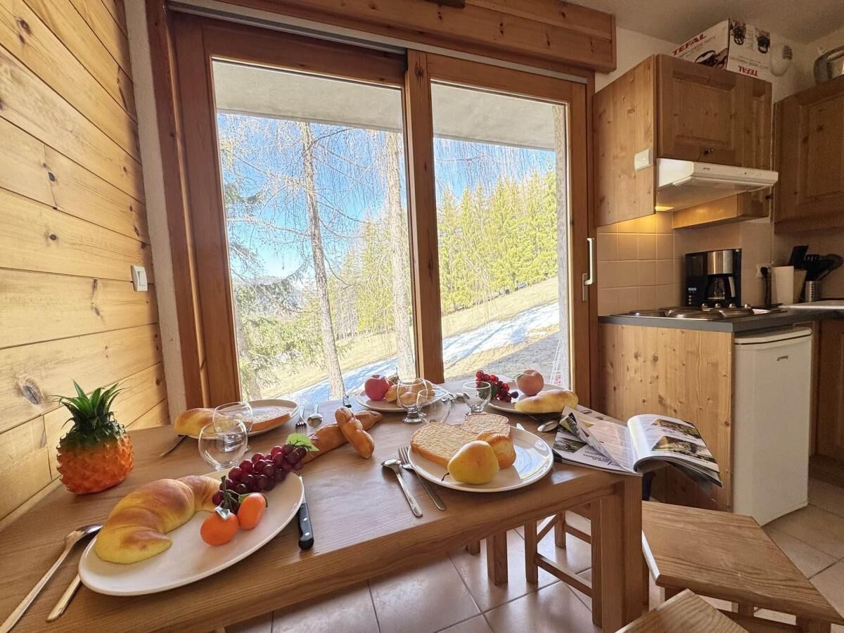 A wooden dining table with breakfast items, a kitchen area with appliances, and a view through large windows.