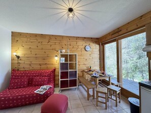 Una habitación acogedora con un muro de madera, un sofá rojo, una mesa de madera con sillas, un estante pequeño y una ventana con vista a árboles.