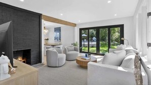 A modern living room with a fireplace, comfortable seating, and a large window offering a view of the outdoors. - Renovated Retreat with Heated Pool (Redding)