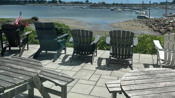 A waterfront patio with Adirondack chairs and a wooden table, overlooking a marina with boats. - Coastal modern 2-bedroom condo in lovely Wells with water views (Wells)