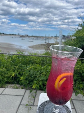 A clear glass with a straw, containing a red beverage and an orange slice, placed on a stone ledge with a view of a body of water and greenery in the background.