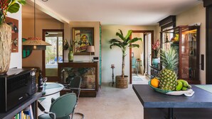 A modern kitchen with a dining area, featuring a central island, a refrigerator, and a variety of plants and fruits.