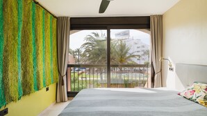 A hotel room with a large bed, a balcony with a view of palm trees and buildings, and a ceiling fan.