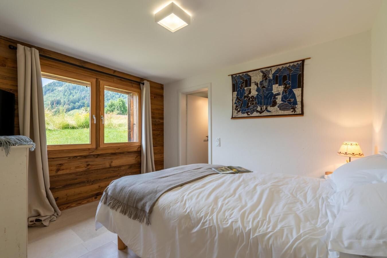 Une chambre avec un plafond en bois, une grande fenêtre, un lit avec une literie blanche, une lampe fixée au mur et une œuvre d’art encadrée.