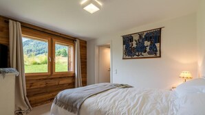 Un dormitorio con techo de madera, un ventanal grande, una cama con ropa de cama blanca, una lámpara de pared y una obra de arte enmarcada.