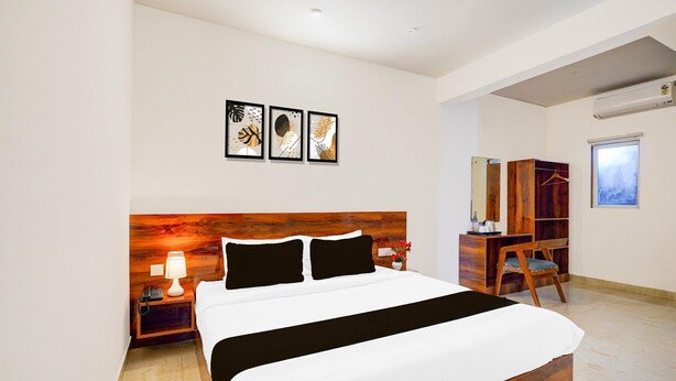 Quarto de hotel com cabeceira de madeira, cama com roupa de cama branca, mesa de cabeceira com abajur, escrivaninha com cadeira e obras de arte na parede.