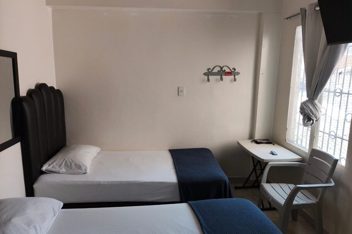 Habitación de hotel con dos camas individuales, una mesita, una silla, televisión y ventana con cortinas.
