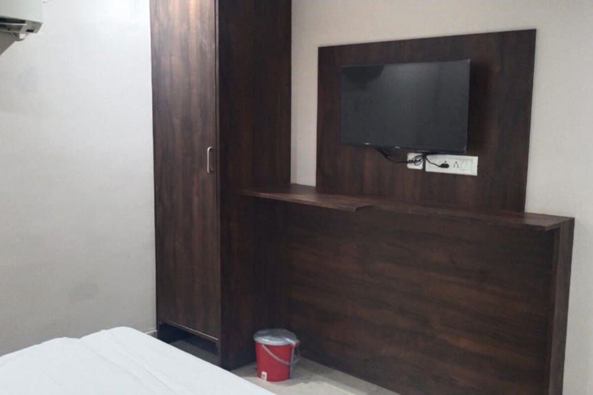 A small room with a bed, a wooden wardrobe, a television mounted on the wall, and a red can on the floor.