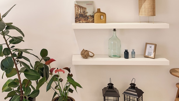Una habitación con estanterías blancas que exhiben libros, velas y objetos decorativos. Hay plantas en macetas y dos faroles en el suelo.