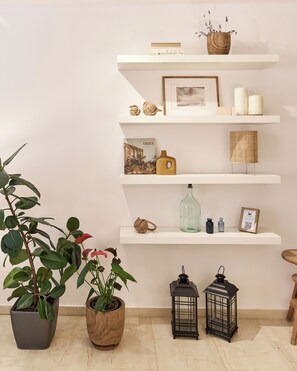 A room with white shelves displaying books, candles, and decorative items. There are potted plants and two lanterns on the floor. - Escape to Kavourify, a luxurious 50 sq.m. located 2-minute from Kavouri Beach! (Vouliagmeni)
